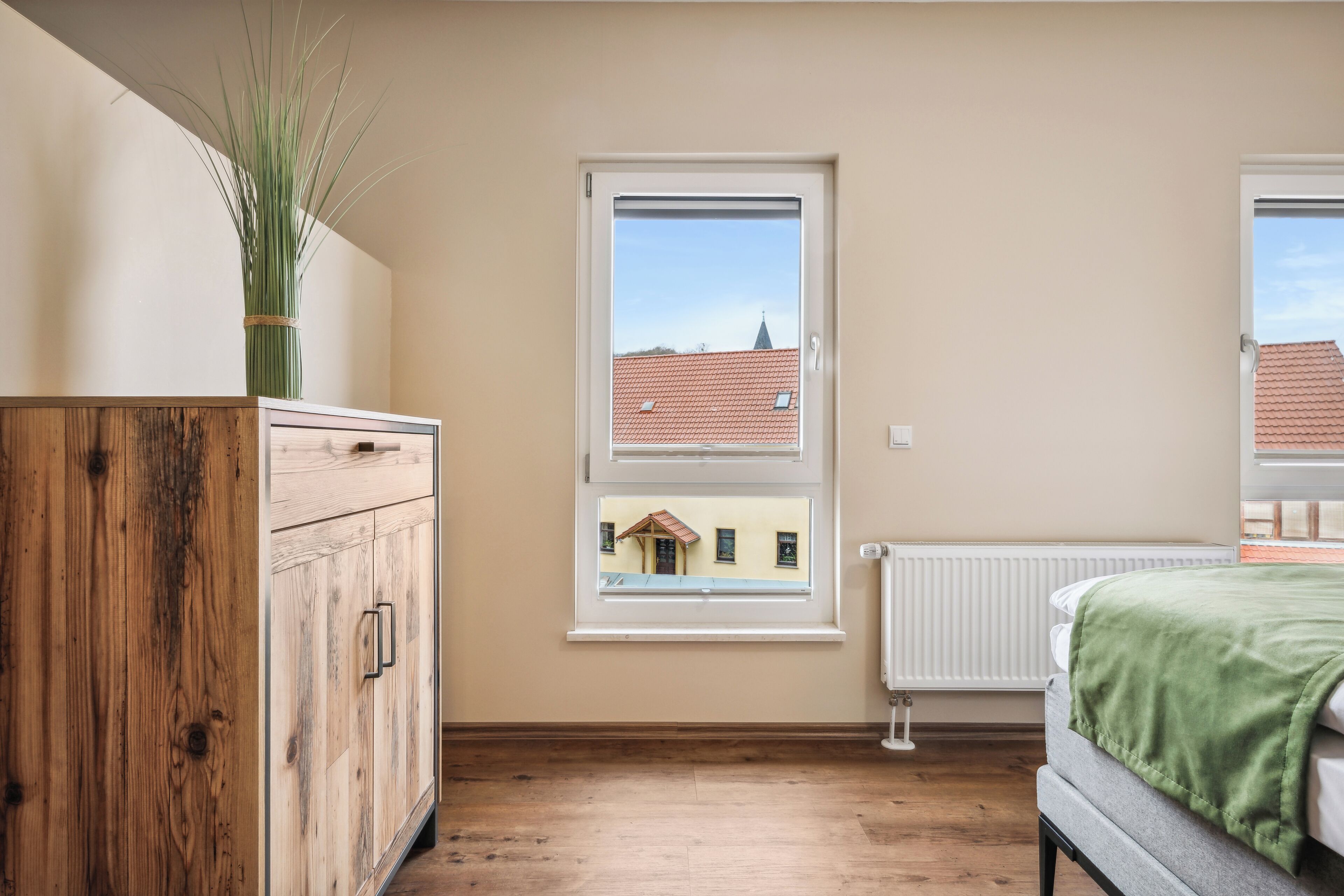 A bedroom with a wooden wardrobe, a radiator, a window with a view of buildings, and a green blanket on the bed.