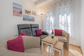A living room with a leather sofa, a woven armchair, a small wooden table with a bottle and glasses, and a large window with sheer curtains. - Moderne Ferienwohnung mit Gartenblick in Koserow (Koserow)