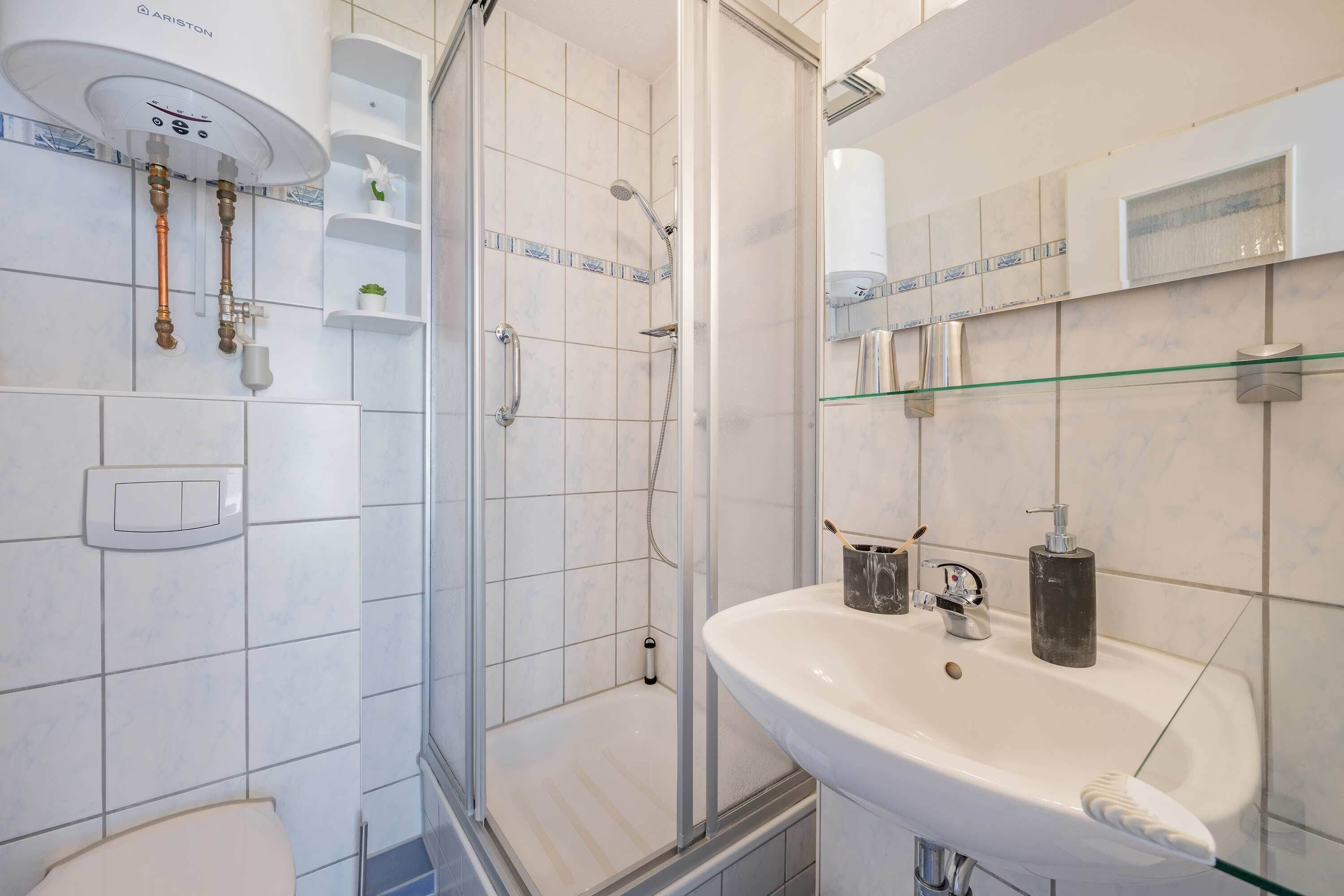 Moderni kylpyhuone, jossa on lasinen suihkuseinä, valkoinen pesuallas, jonka alla on lasihylly, sekä wc-istuin.