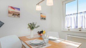 Un coin repas avec une table en bois, des chaises blanches, un vase avec des plantes, un saladier de fruits et une fenêtre avec des rideaux.