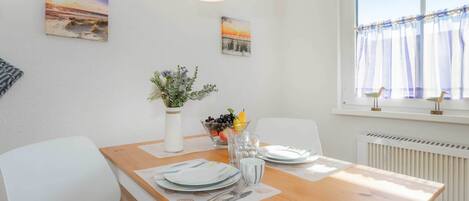 A dining area with a wooden table, white chairs, a vase with plants, a bowl of fruit, and a window with curtains.