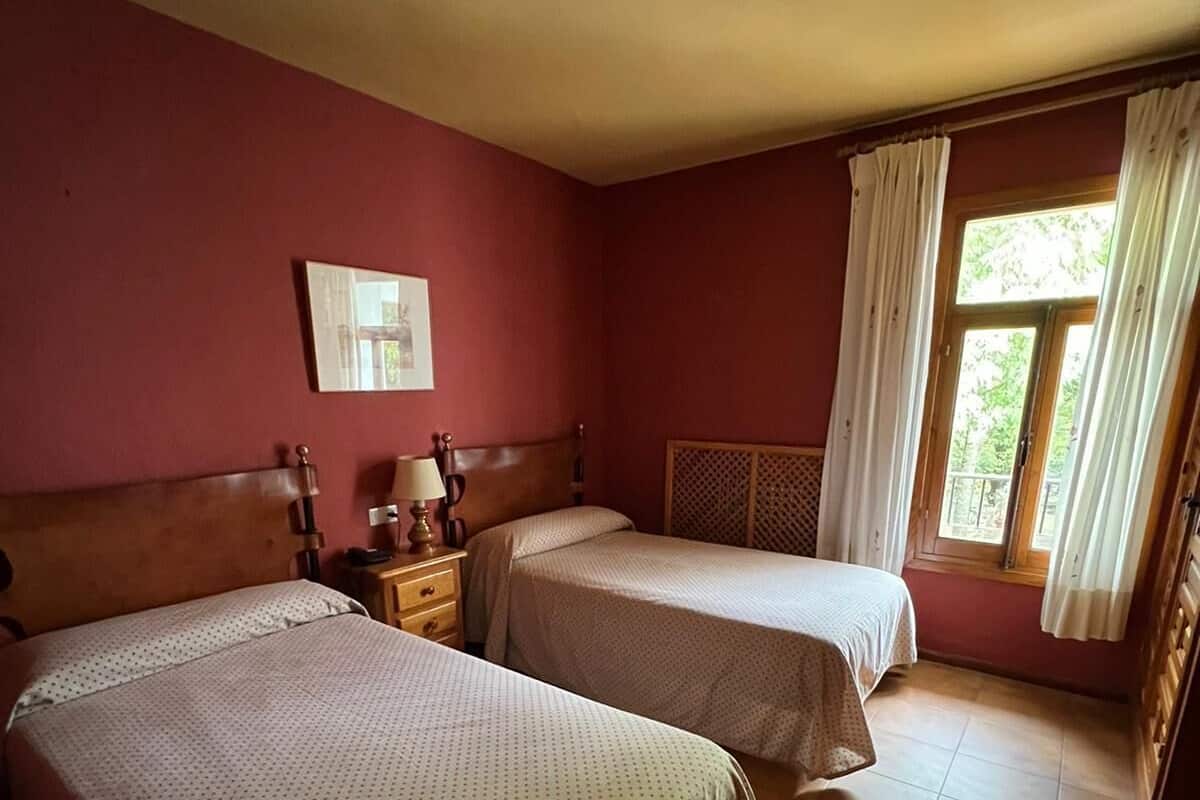 Ein Zimmer mit zwei Betten, einem Nachttisch mit Lampe, einem Fenster mit Vorhängen und einem Bild an der Wand.