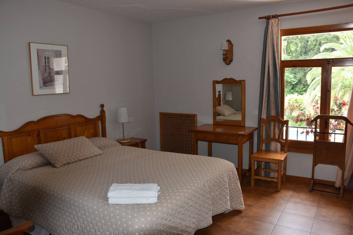 Ein Schlafzimmer mit Bett, Schreibtisch, Stuhl und einem Fenster mit Blick auf Grünflächen.