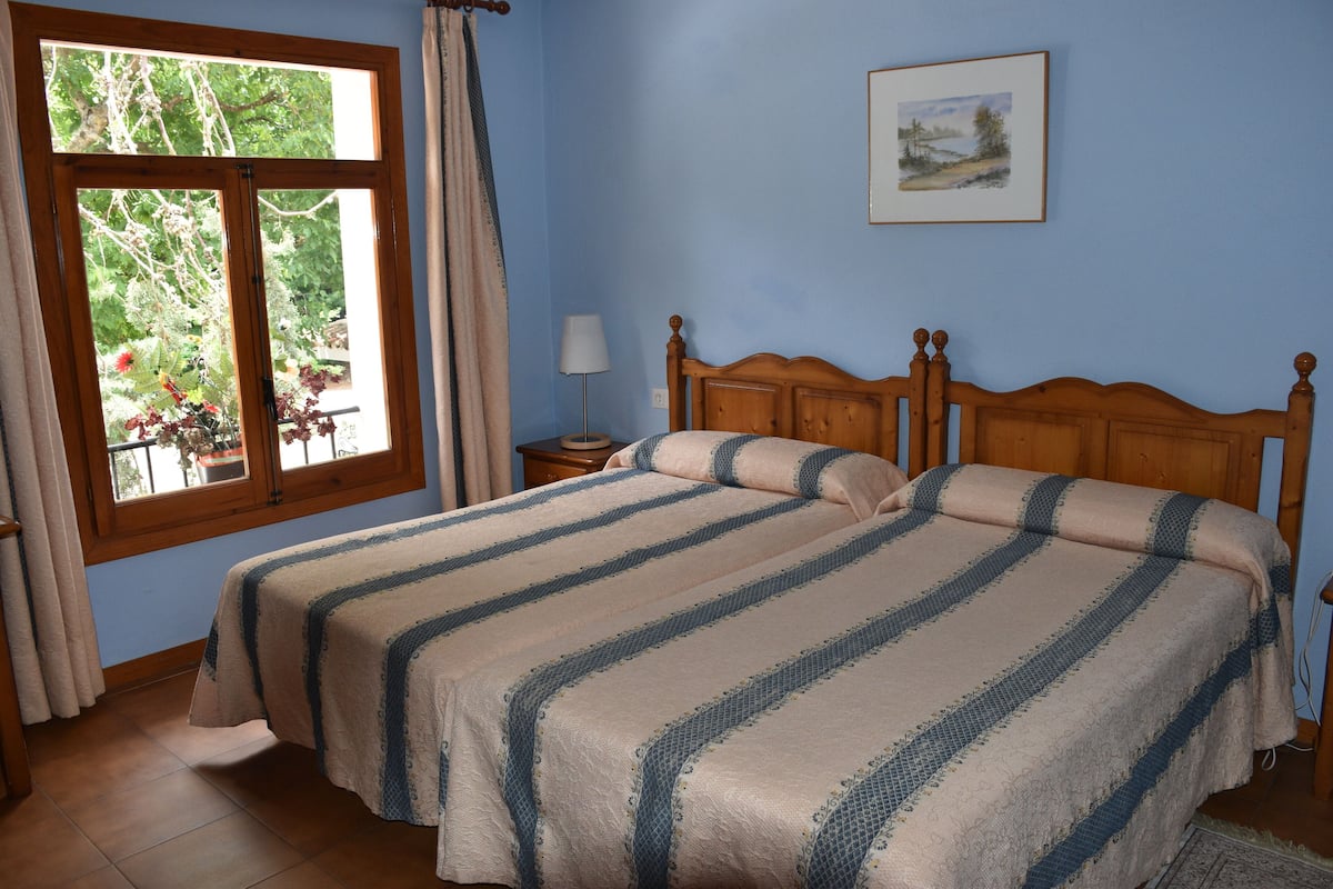 Ein Schlafzimmer mit zwei Betten, einem Fenster mit Vorhängen, einem Nachttisch mit Lampe und einem gerahmten Bild an der Wand.