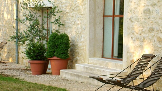 A stone building with a window, two potted plants, and two lounge chairs.