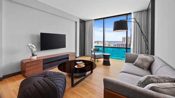A modern living room with a flat-screen TV, a round coffee table, a sofa, and a view of the city and water. - Marriott Executive Apartments Port Moresby (Port Moresby)