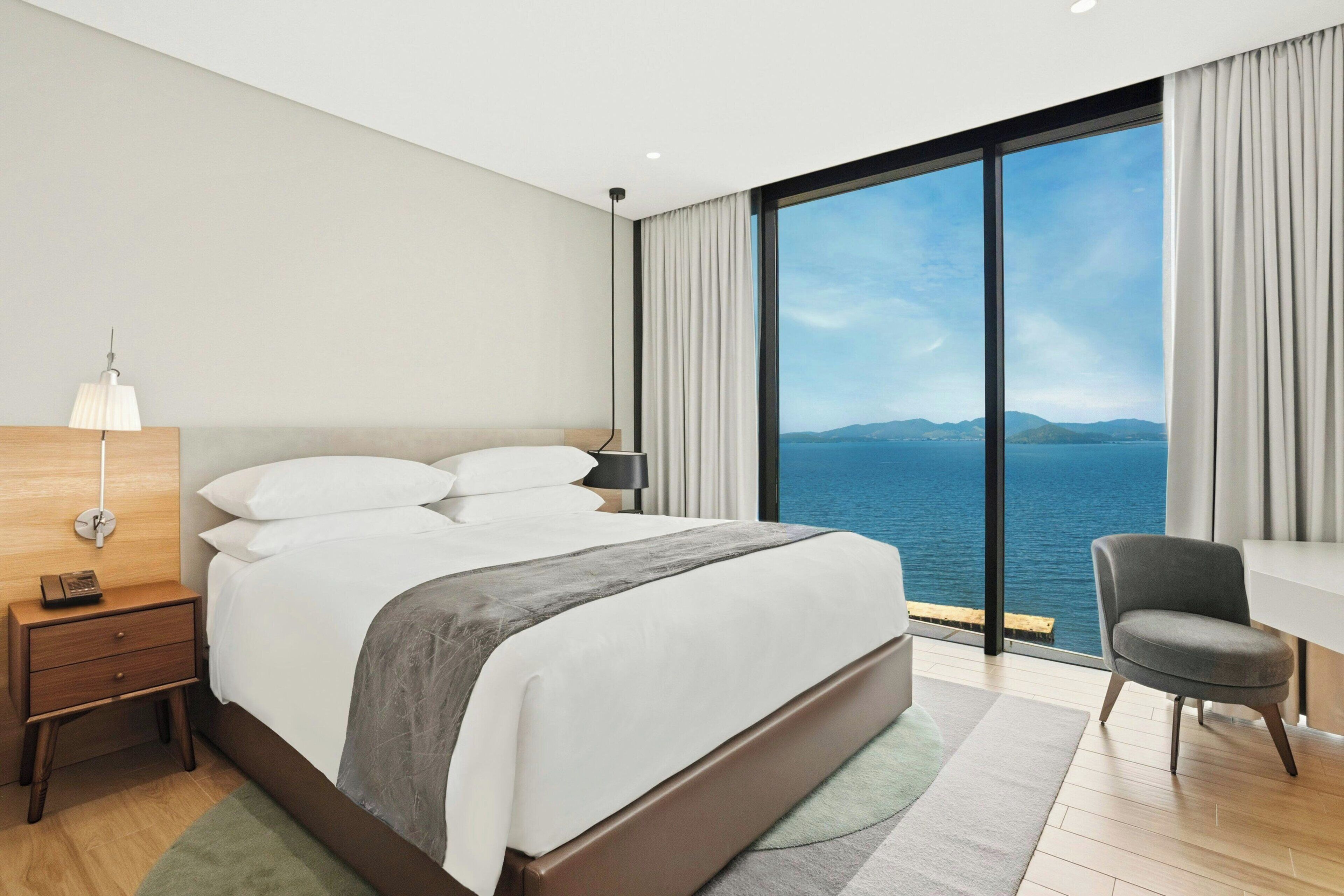 A modern hotel room with a large bed, a desk, and a view of the sea.