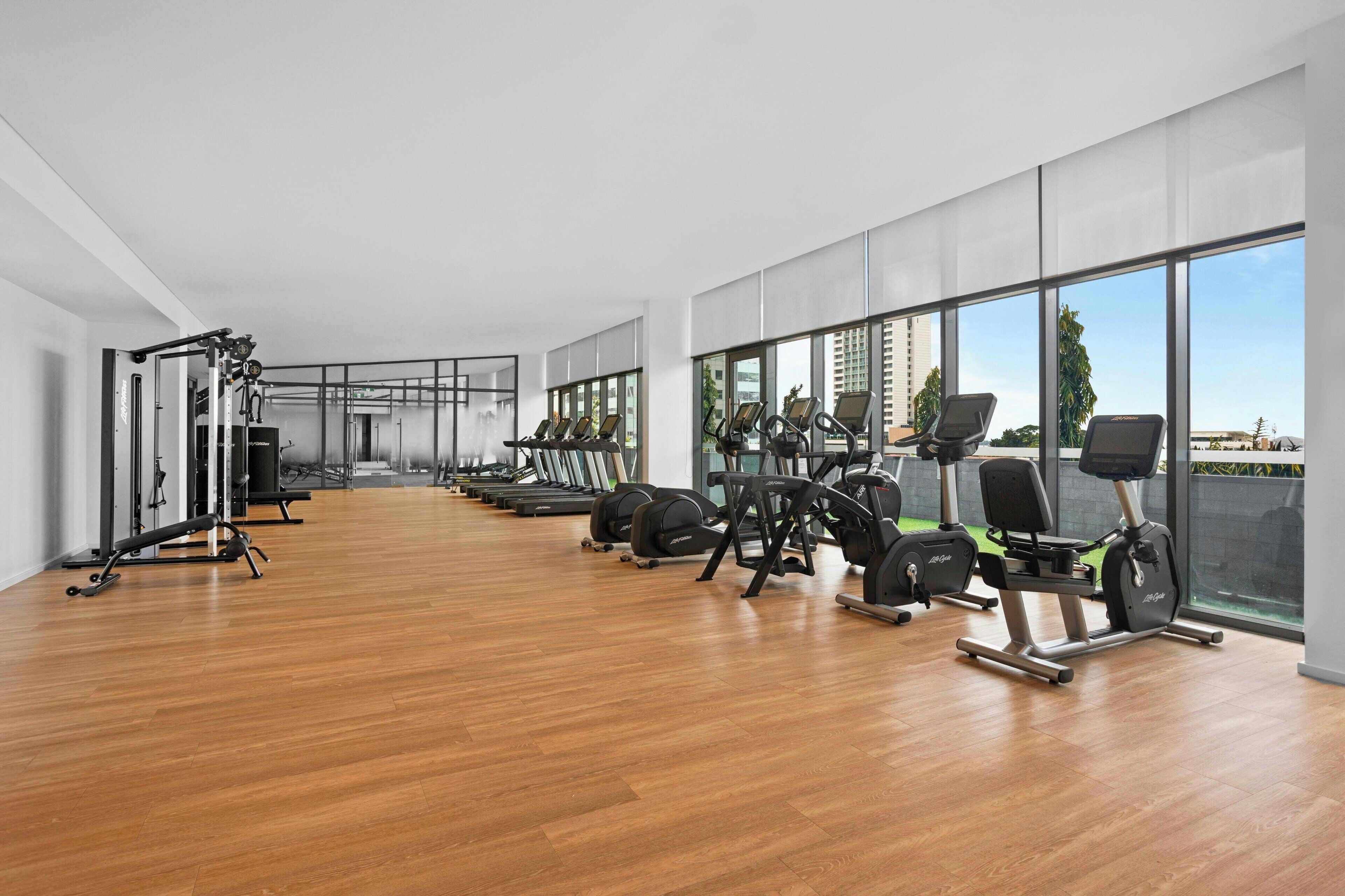 A modern gym with cardio machines, weight stations, and a large window offering a city view.