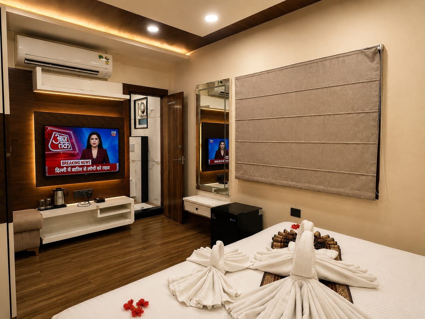 A hotel room with a TV displaying a news channel, a minibar, a bed with towels, and a mirror.