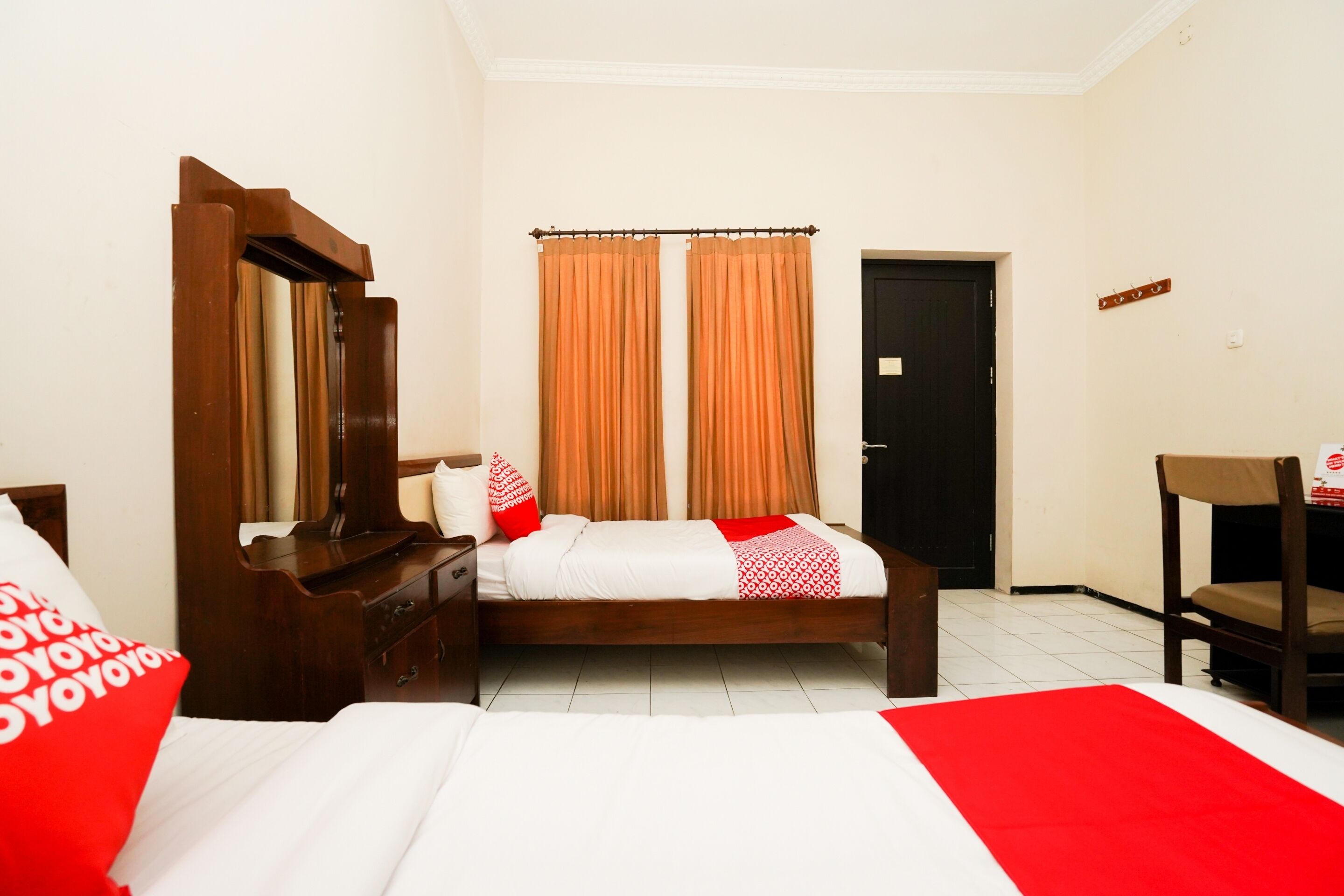 A hotel room with a bed, a wooden dresser, a mirror, orange curtains, and a door.