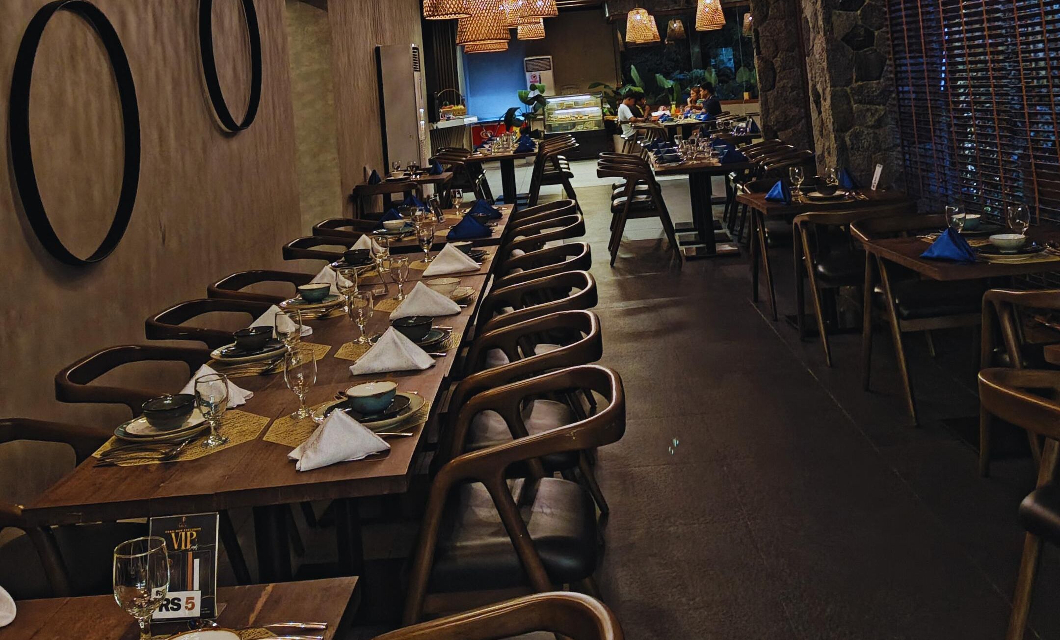 A long dining table set with multiple chairs, a buffet area in the background, and a stone wall with a grid-like pattern.