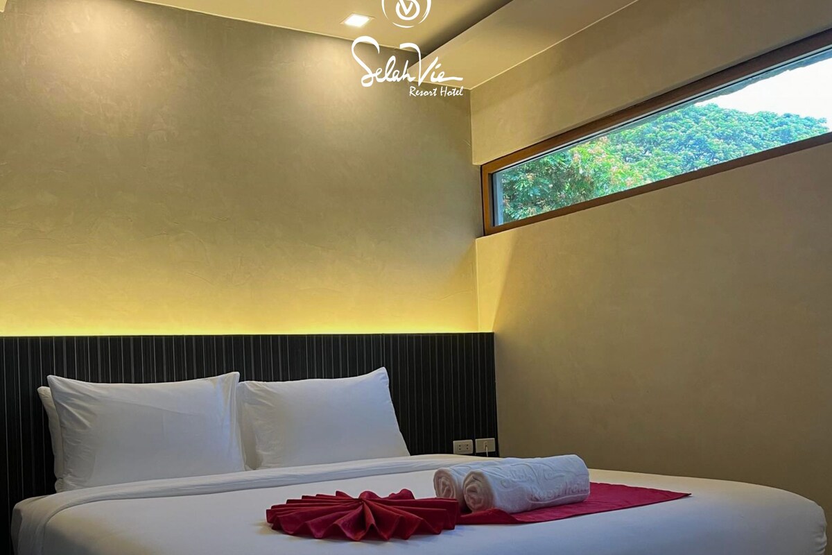 A modern hotel room with a large bed, white linens, and a red towel. The room has a view of greenery through a window.