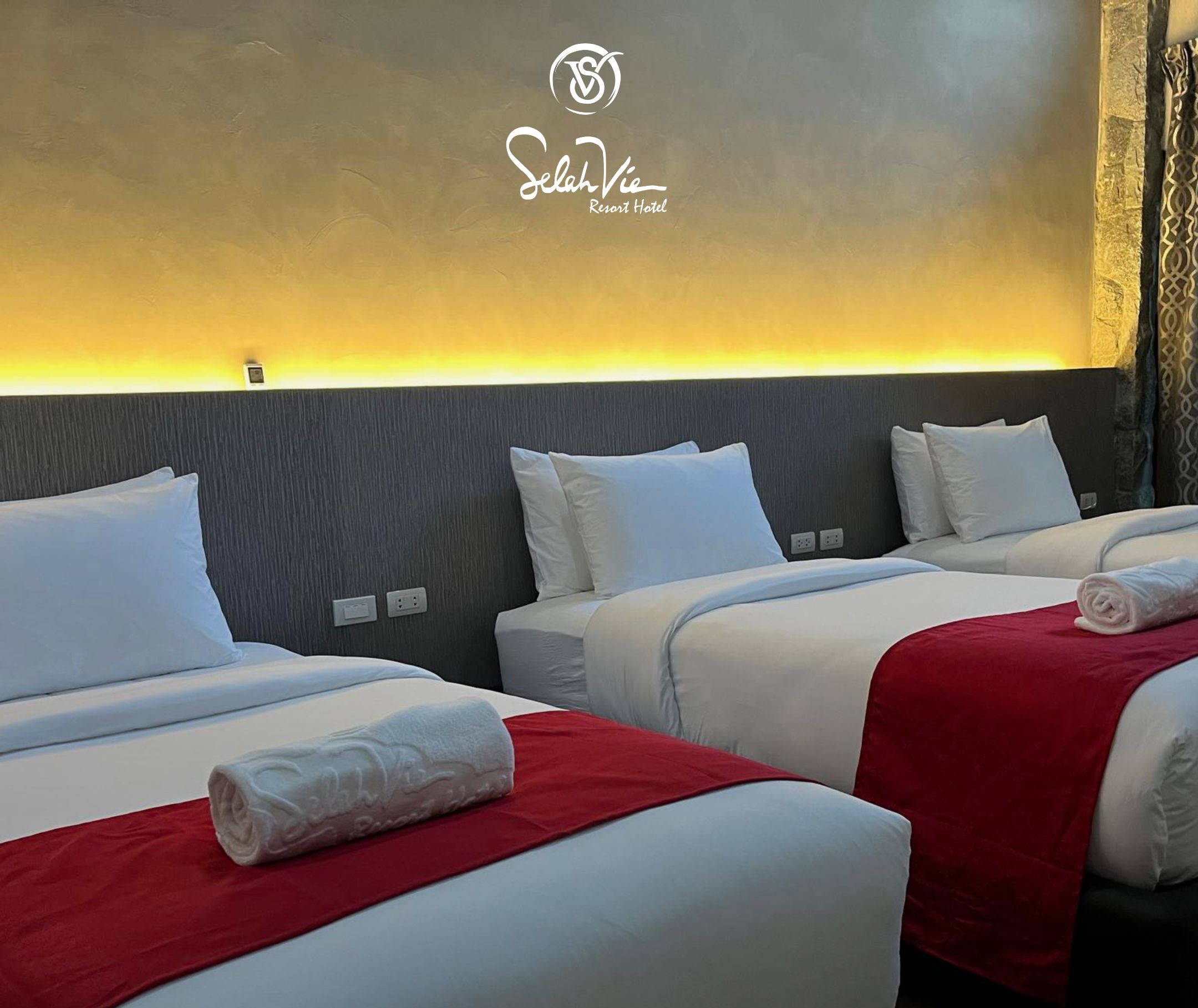 A hotel room with two beds, each with white bedding and red blankets. The room has a modern design with a grey headboard and a wall-mounted light fixture. The hotel's name, Selah Vie Resort Hotel, is displayed on the wall.