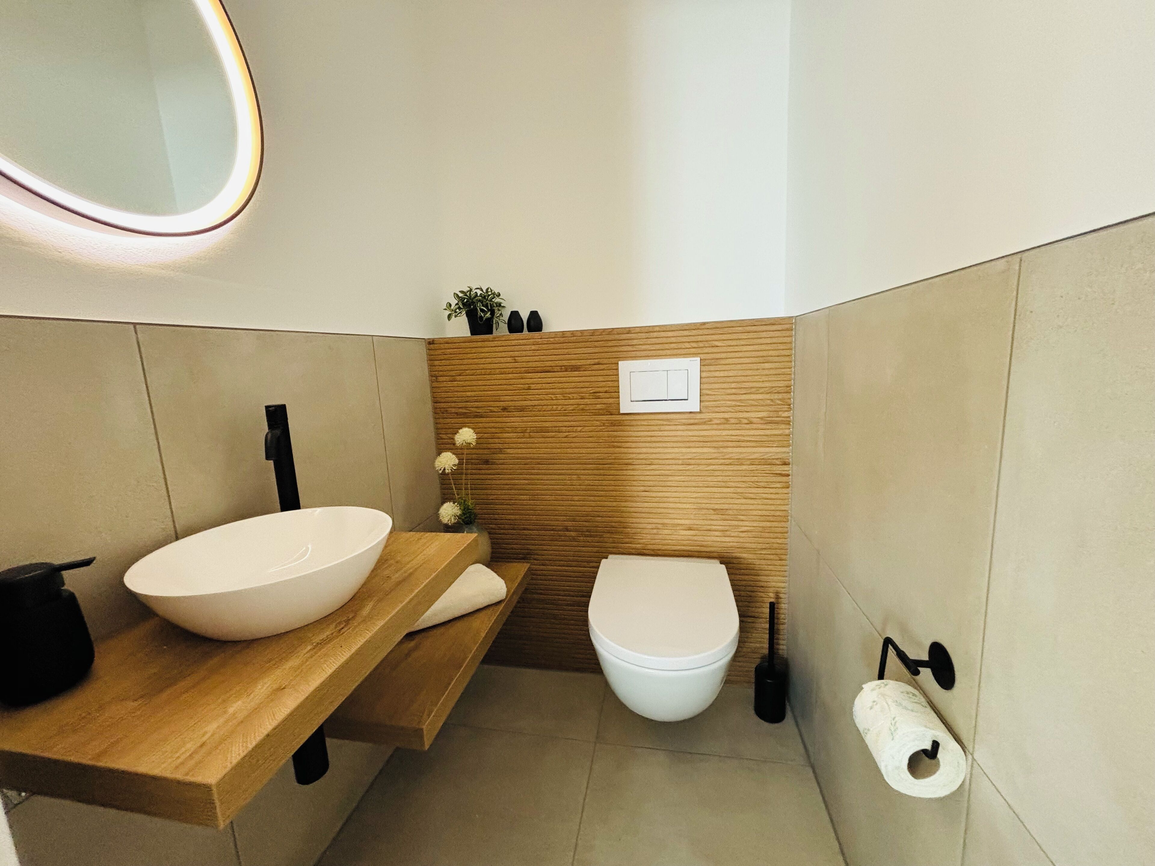 A modern bathroom with a wooden vanity, a large white basin, a toilet, and a wall-mounted toilet paper holder.