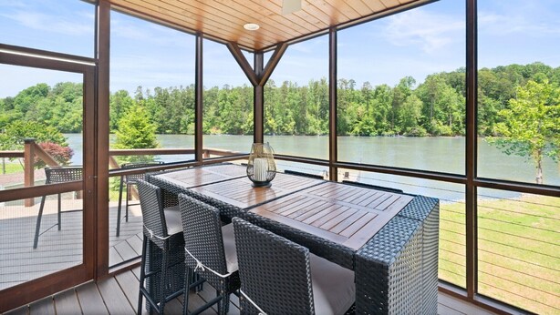 A spacious balcony with a wooden table and chairs, large windows offering a view of a lake and forest.