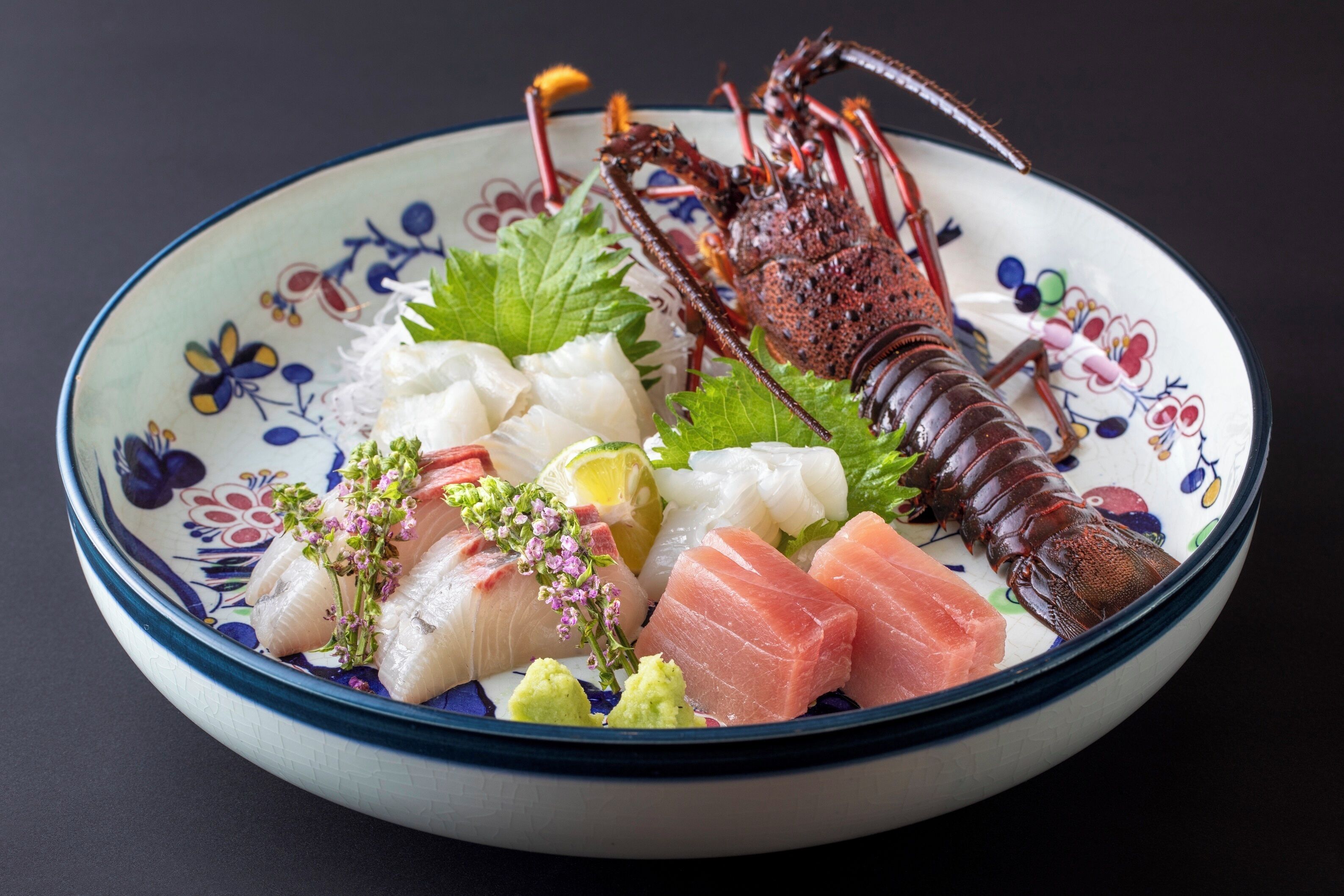A bowl of sashimi with a lobster tail, garnished with herbs and lemon.