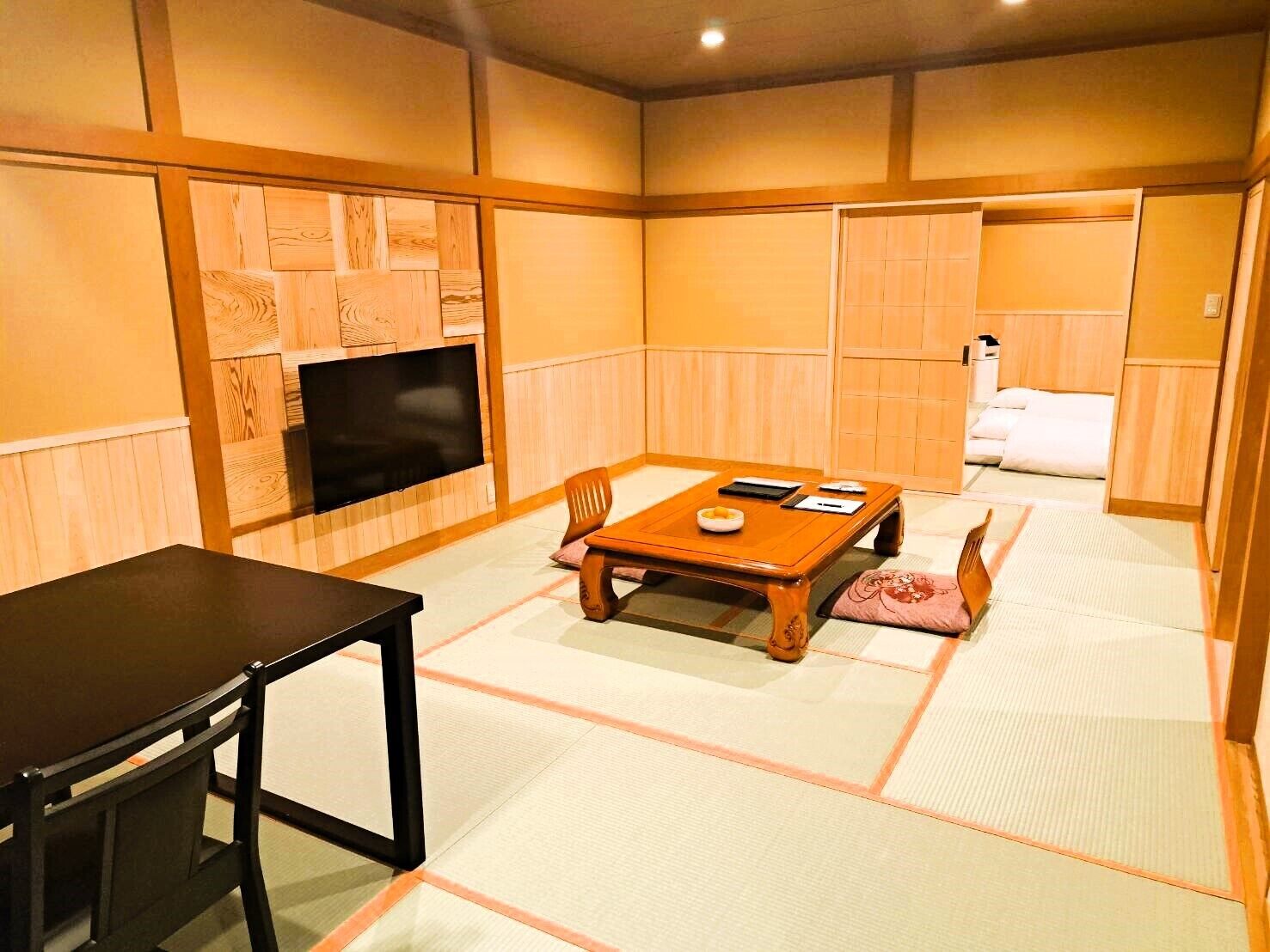 Una habitación tradicional japonesa con suelo de tatami, una mesa de madera baja, un televisor montado en la pared y una cama con manta blanca.
