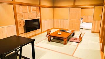 Una habitación tradicional japonesa con suelo de tatami, una mesa de madera baja, un televisor montado en la pared y una cama con manta blanca.