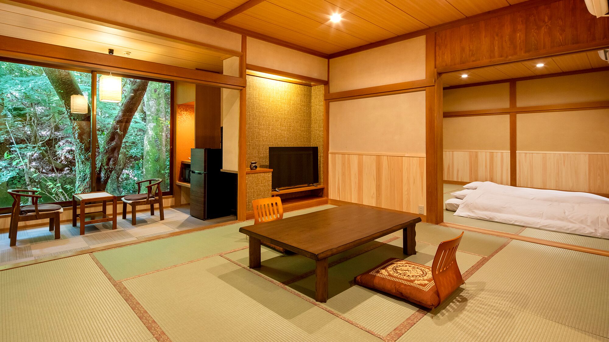 Una habitación tradicional japonesa con suelo de tatami, una mesa baja, sillas, un televisor y vistas a un bosque a través de amplios ventanales.