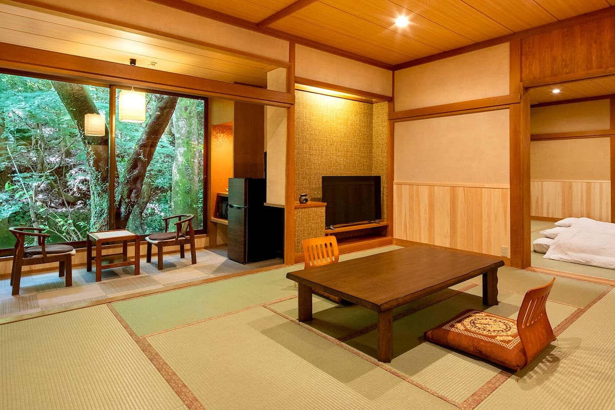 A traditional Japanese-style room with tatami flooring, a low table, chairs, a television, and a view of a forest through large windows.