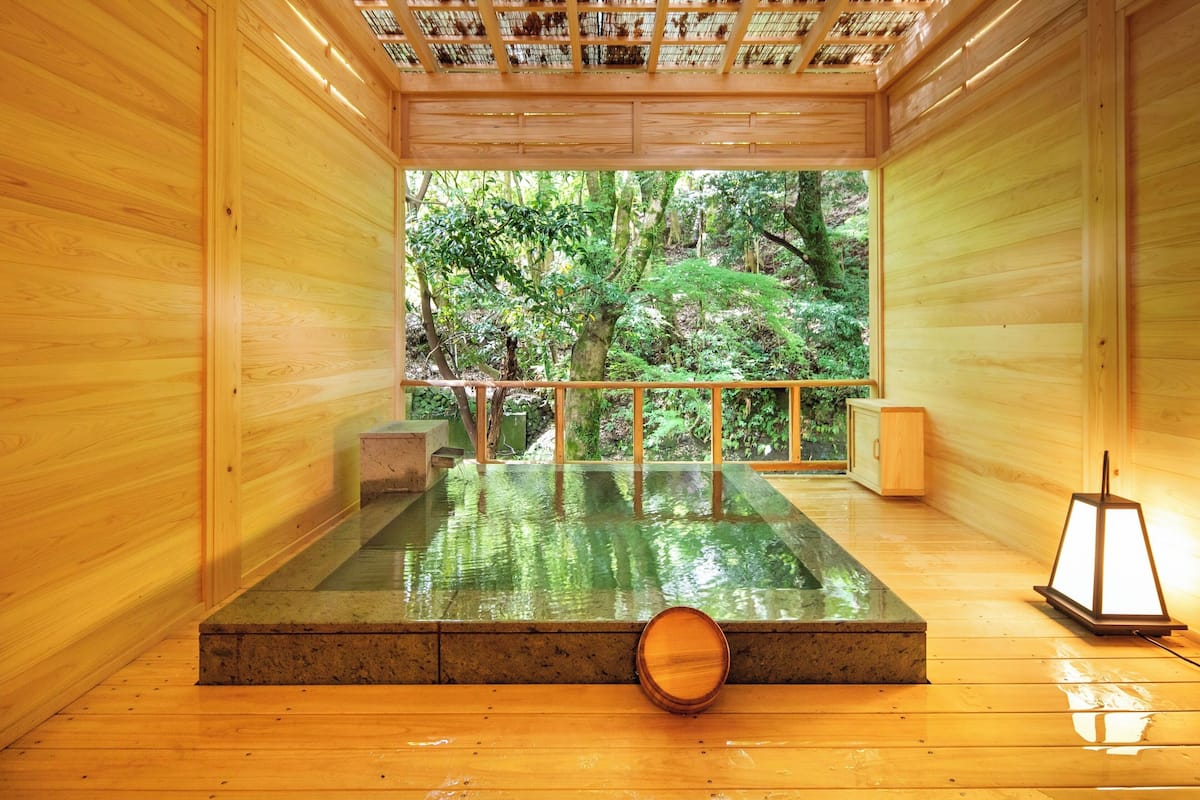 A wooden interior with a glass-enclosed hot spring area, wooden flooring, and a view of a forested area.