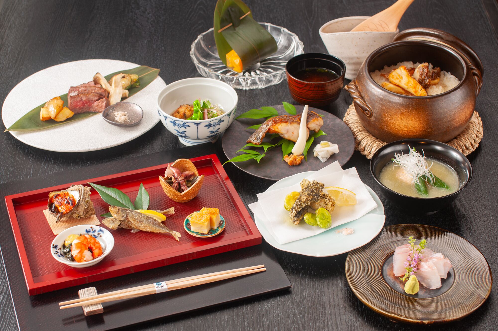 A variety of Japanese dishes including sushi, sashimi, and a hot pot, arranged on a dark table.