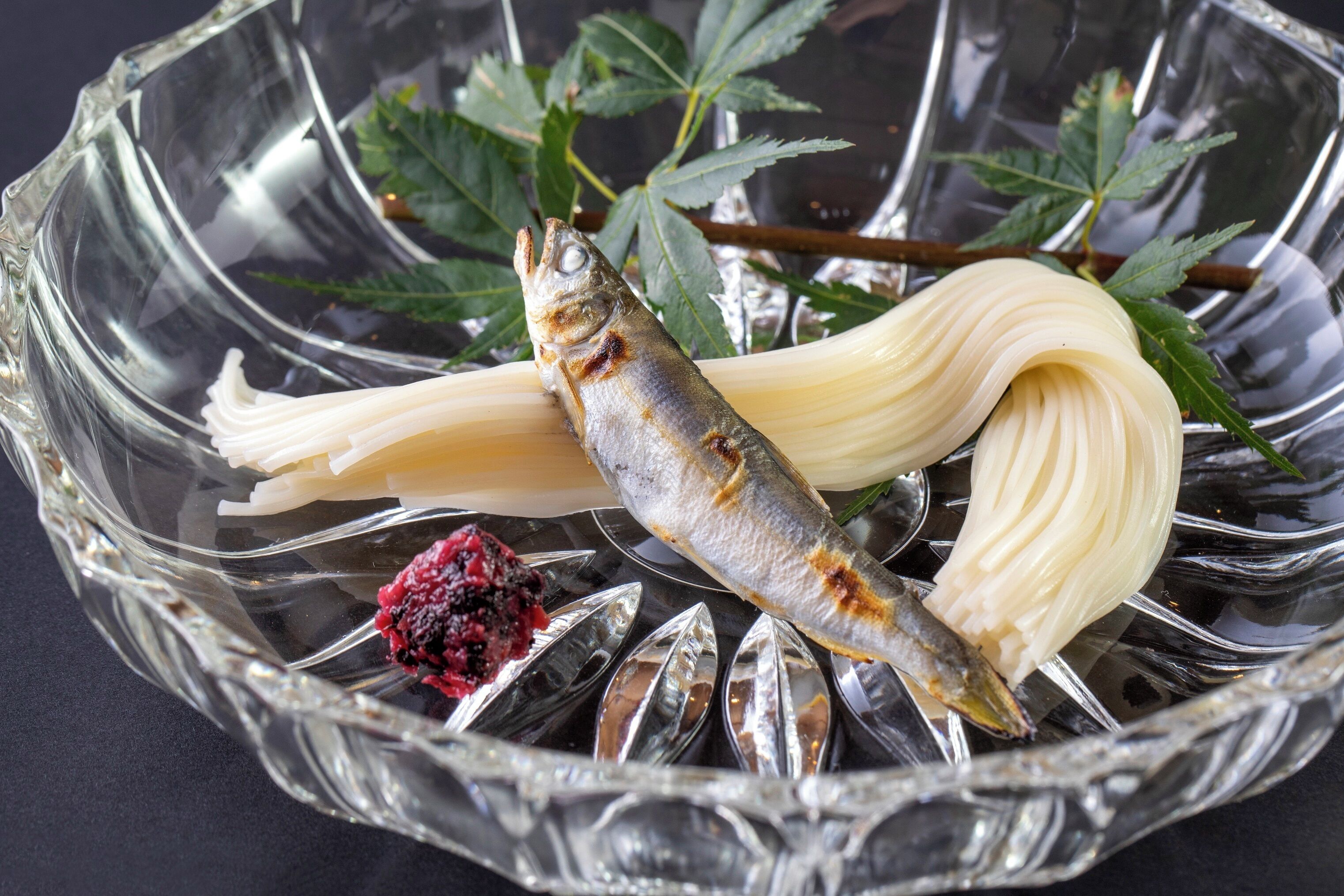 A glass bowl containing a fish, noodles, and a berry.