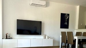 Een moderne woonkamer met een flatscreen televisie, witte kasten, een eethoek met stoelen en een witte salontafel.