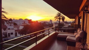 Een balkon met uitzicht op een zonsondergang boven een stadsbeeld, een bank en een bloempot.