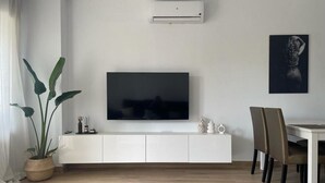 Een moderne woonkamer met een flatscreen televisie, witte kasten, een eettafel met stoelen en een grote plant.