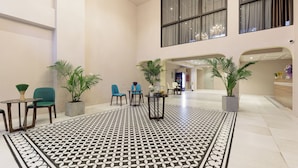 A spacious lobby with a patterned floor, seating areas, potted plants, and large windows.