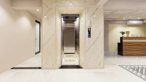 An elevator with marble walls and a patterned floor leading to a reception area with a wooden desk and a potted plant.