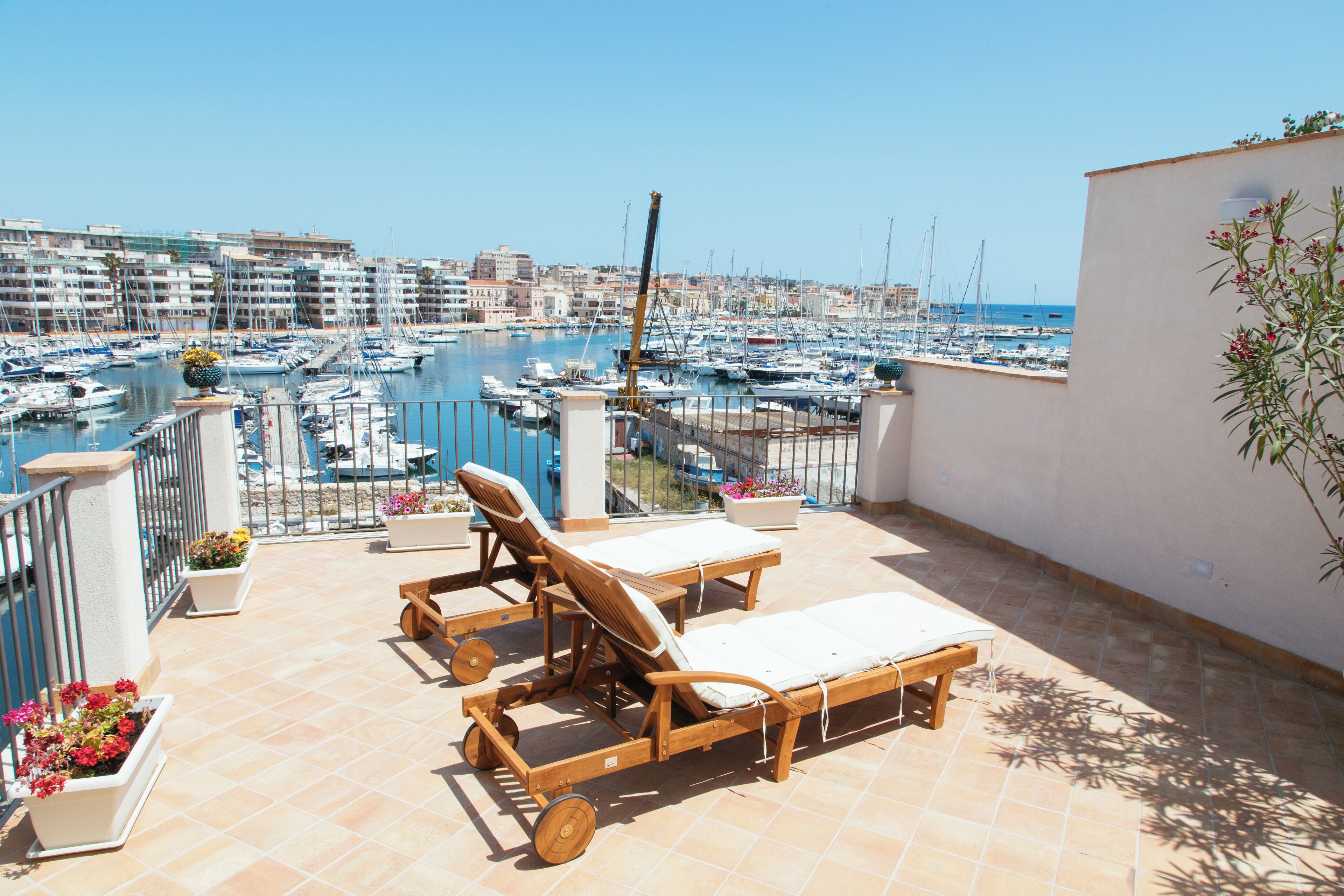 A terrace with two lounge chairs and a table, overlooking a marina with boats and buildings in the background.
