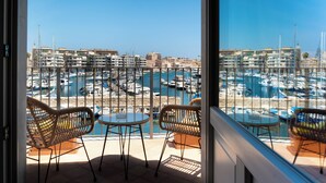Uma varanda com cadeiras de vime e uma pequena mesa, com vista para uma marina com barcos e edifícios.