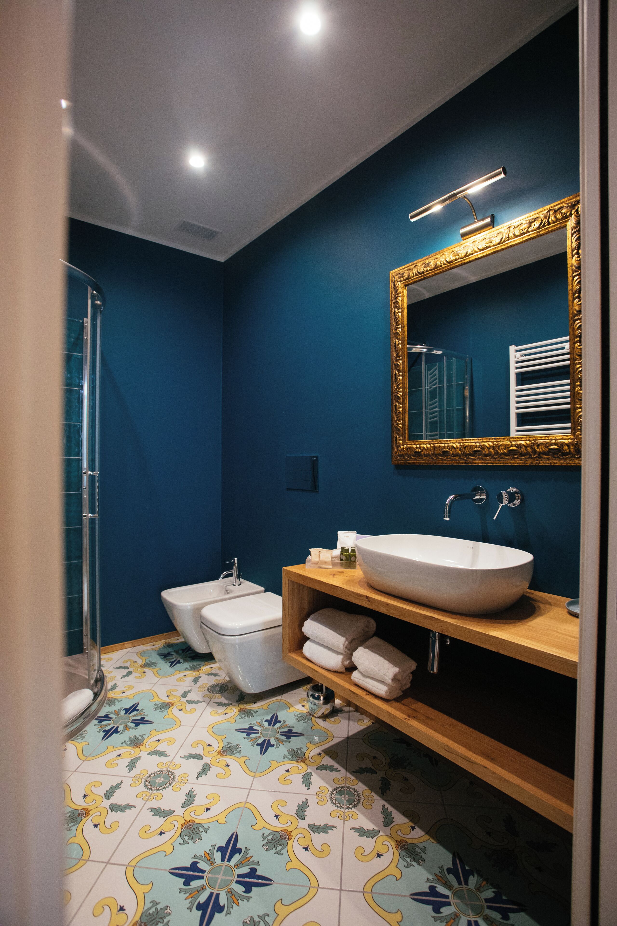 Un baño con un lavabo grande de color blanco, un espejo con marco dorado, una ducha y un suelo de baldosas con estampado floral.