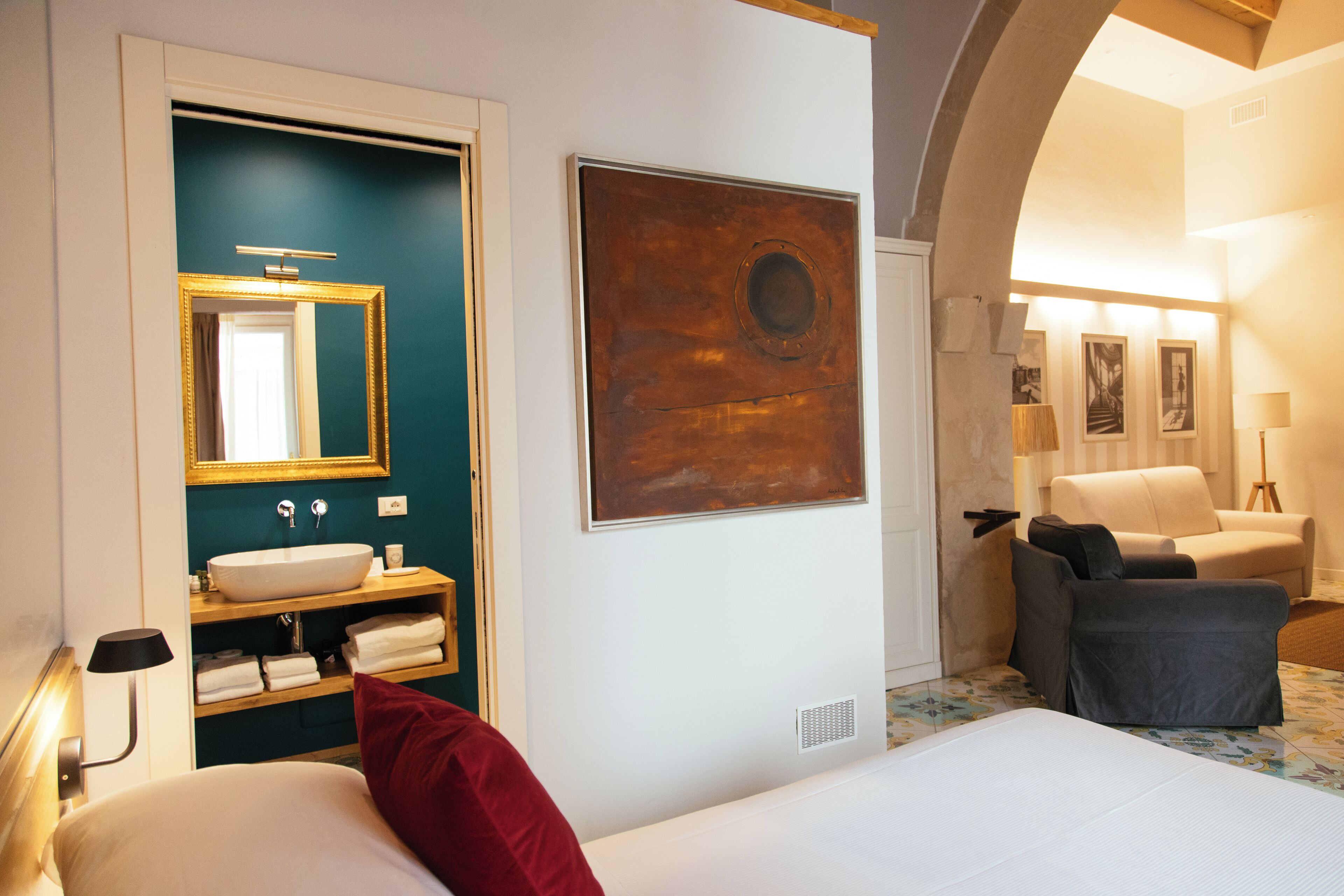 Una habitación de hotel con cama, baño con lavamanos y espejo, un cuadro en la pared y un rincón de estar con sofá y sillones.