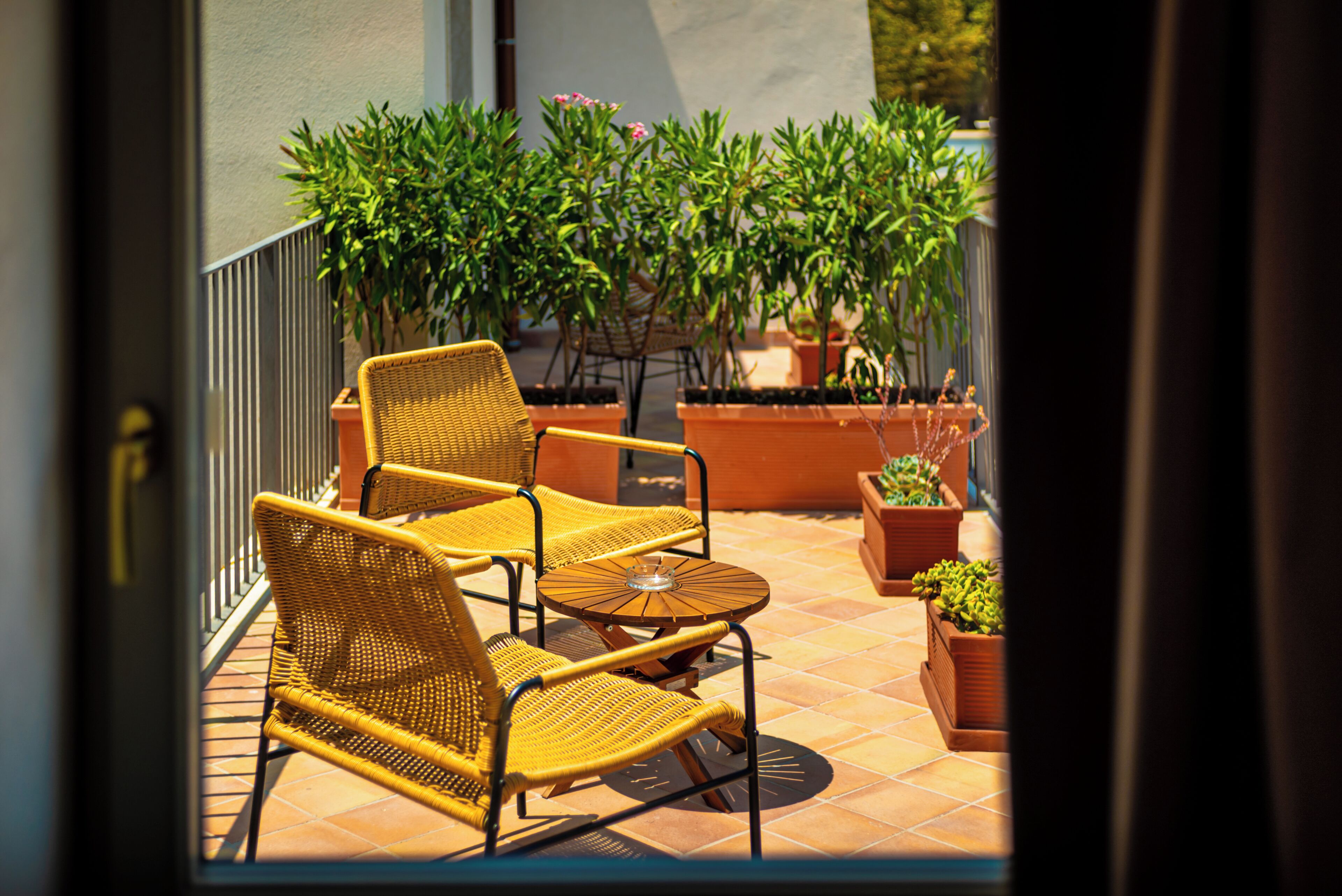 A patio with wicker chairs and a small table, surrounded by potted plants and a tiled floor.