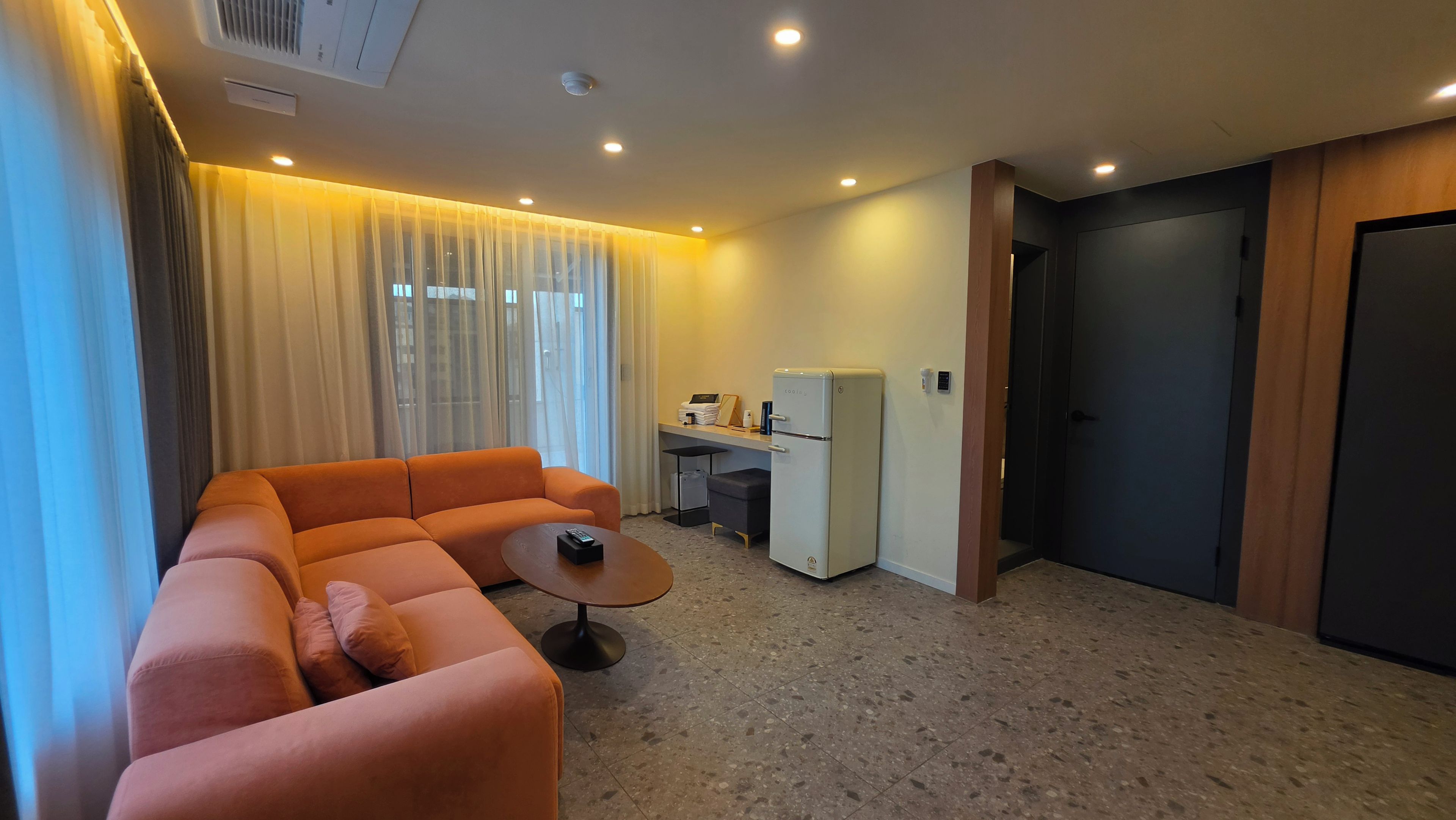 A modern hotel room with a sofa, a small table, a refrigerator, a desk, and a door.