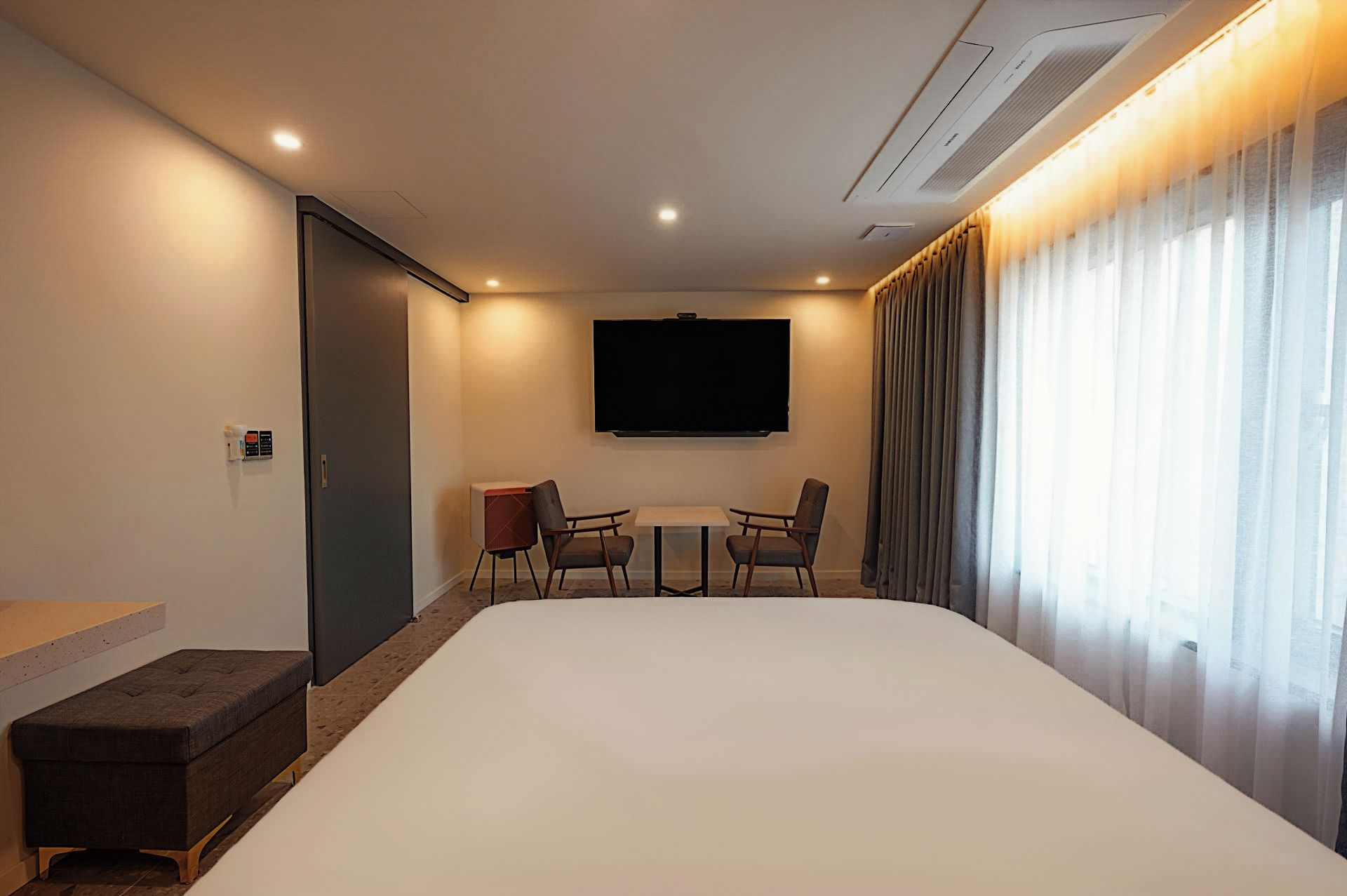 Habitación de hotel con una cama grande, un televisor de pantalla plana en la pared, una pequeña mesa con dos sillas y una ventana con cortinas.