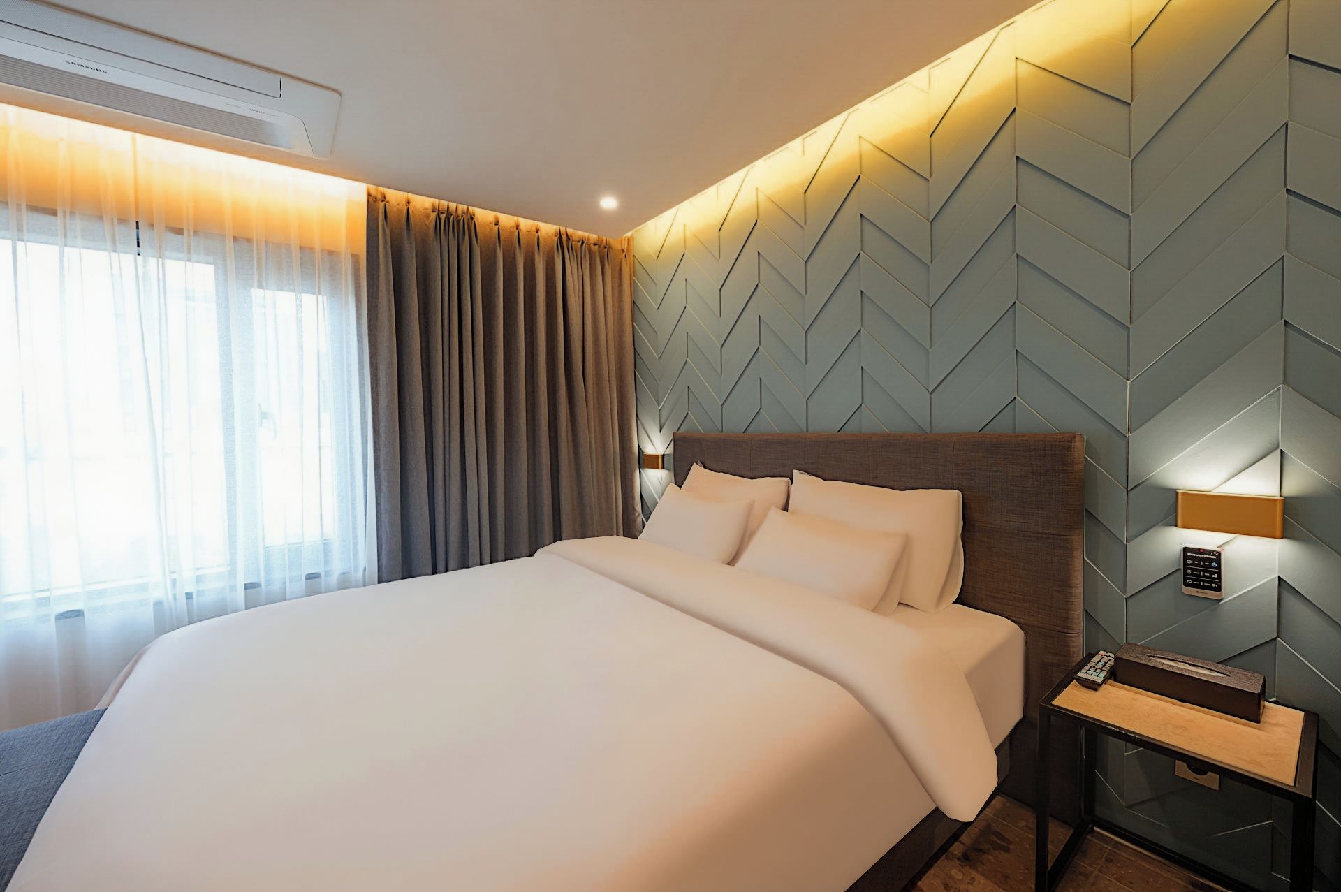 A hotel room with a large bed, a nightstand, a wall-mounted lamp, and a window with curtains.
