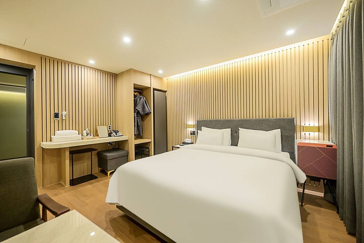 Ein modernes Hotelzimmer mit einem großen Bett, einem Schreibtisch mit Stuhl, einem Kleiderschrank und einem Nachttisch mit Lampe.
