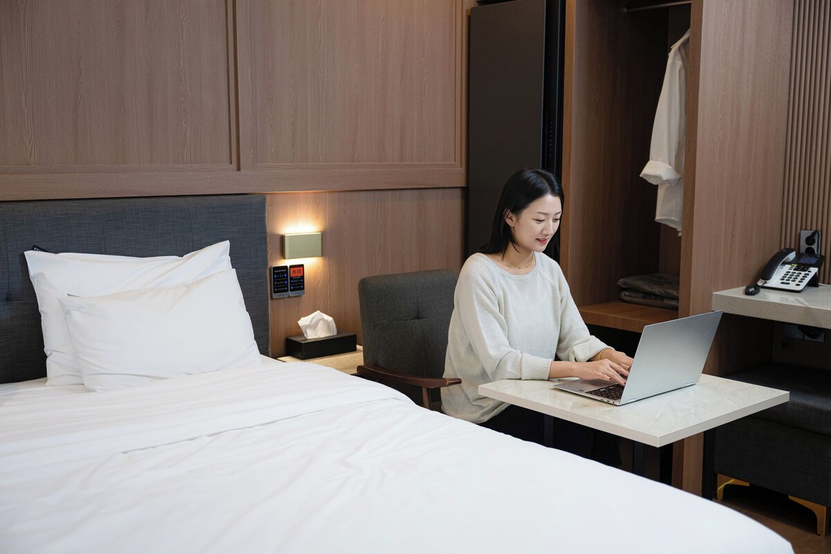 A hotel room with a bed, a desk with a laptop, a telephone, and a nightstand with a lamp.
