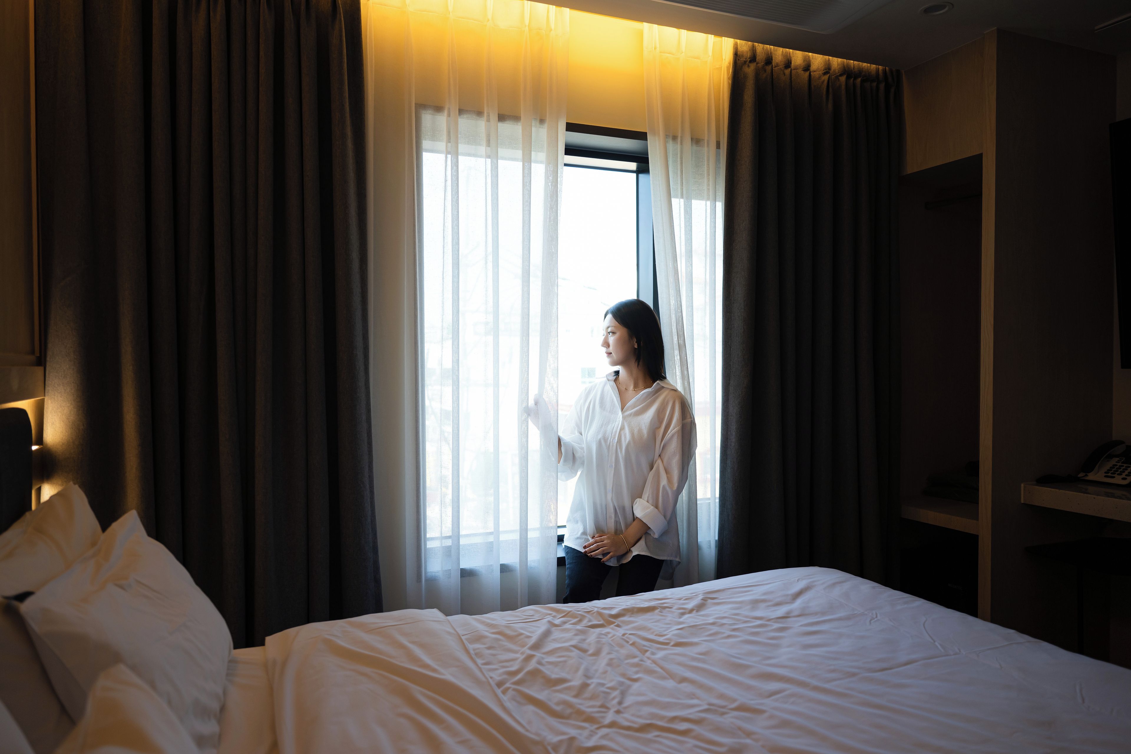 A hotel room with a bed, a window with curtains, and a telephone on a shelf.