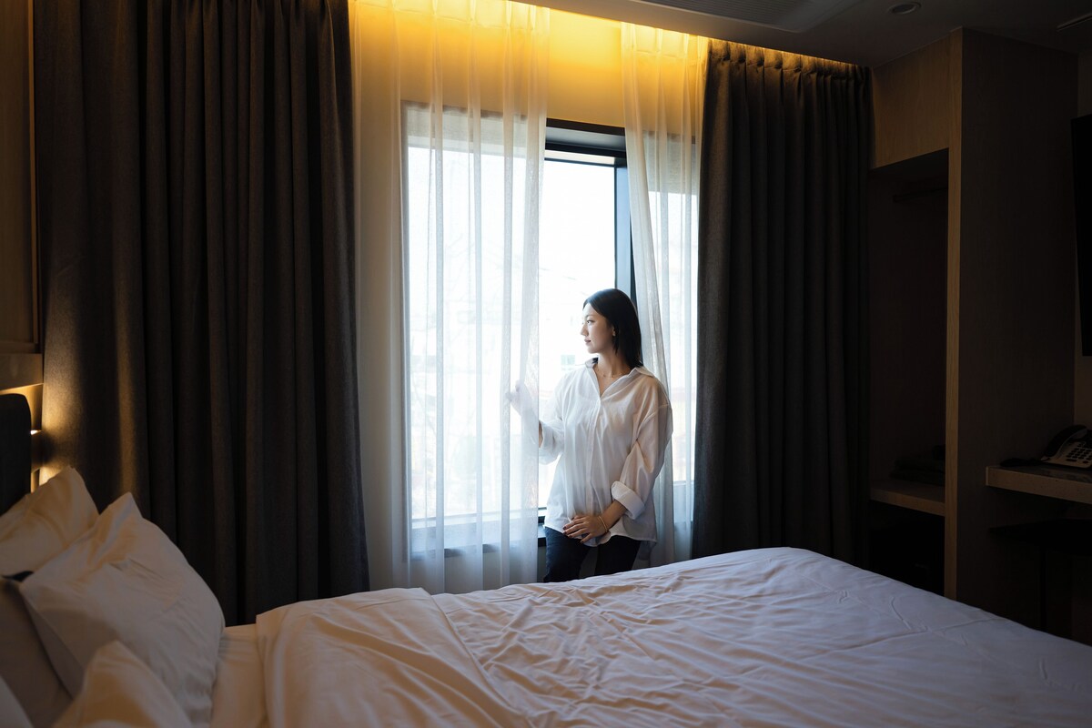 A hotel room with a bed, a window with curtains, and a telephone on a shelf.