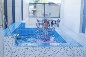 A child in a blue tiled bathtub with a showerhead, surrounded by a tiled floor and a glass railing in the background. - Mokpo H Avenue Hotel Peace Plaza (Mokpo)