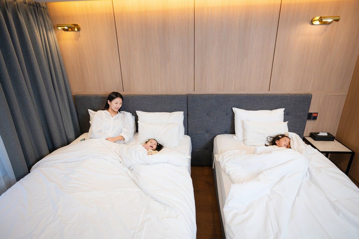 A hotel room with two beds, a nightstand, and a wall-mounted control panel.