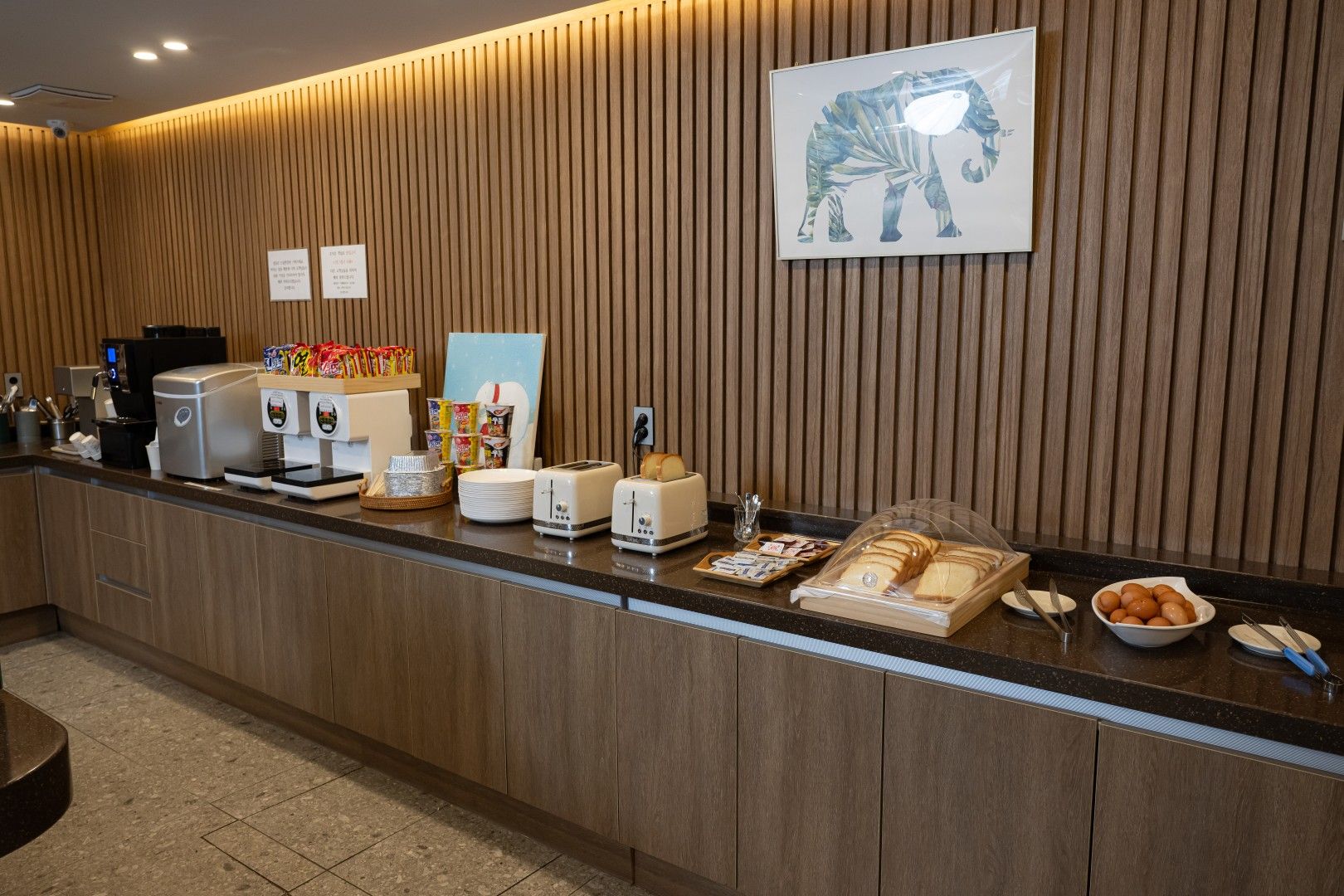 Un espace d’accueil d’hôtel avec un mur recouvert de panneaux en bois, une œuvre d’art représentant un éléphant encadrée, une machine à café, une balance et divers articles pour le petit-déjeuner disposés sur le comptoir.