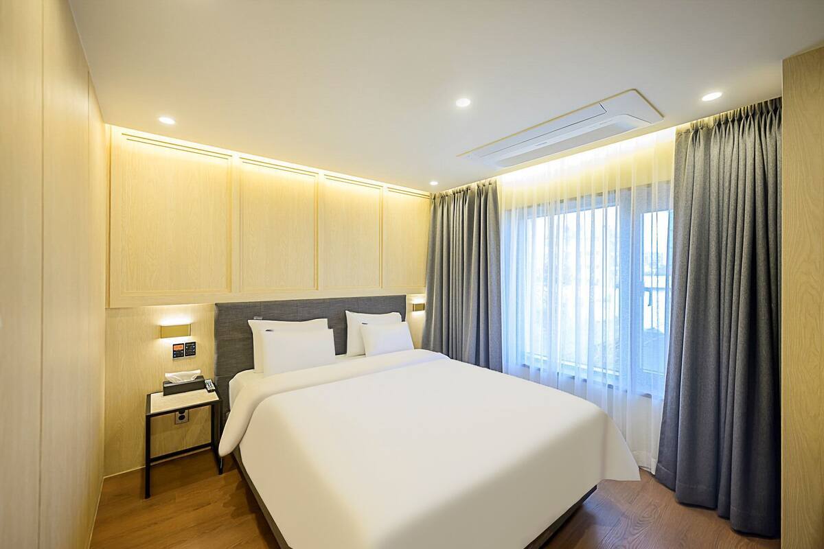 Ein modernes Hotelzimmer mit einem großen Bett, einem Nachttisch mit Lampe und einem Fenster mit Vorhängen.