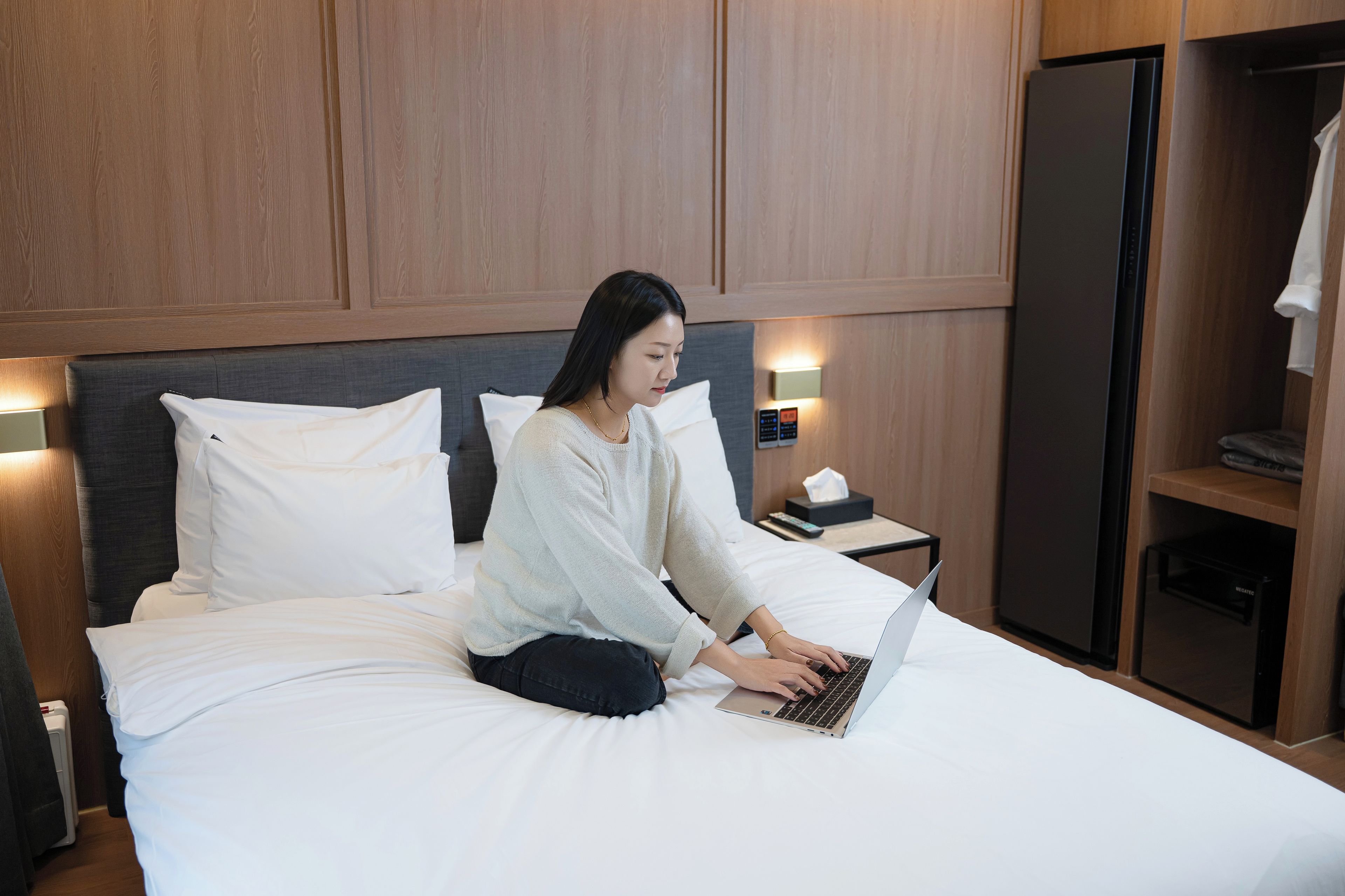 A person sitting on a bed with a laptop, in a room with wooden paneling and a nightstand with a remote control.