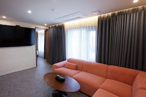 A modern hotel room with a sofa, a coffee table, a flat-screen TV mounted on the wall, and a view through the window. - Mokpo H Avenue Hotel Peace Plaza (Mokpo)